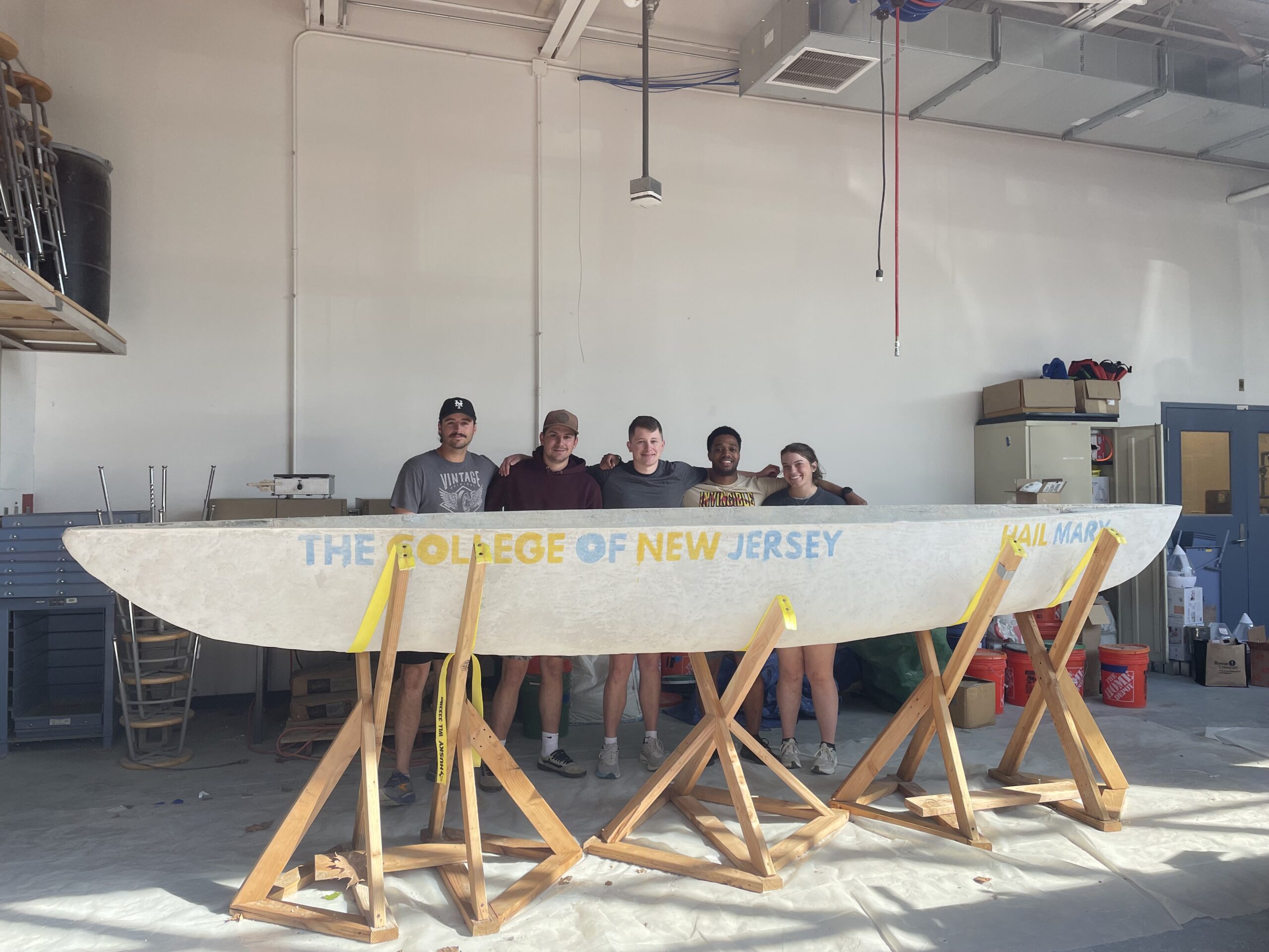 The 2025 Concrete Canoe team standing behind their canoe (The "Hail Mary") while it is on its wooden stands in their laboratory. The canoe has "The College of New Jersey" (left) and "Hail Mary" (right) stained on the side. The color of the words alternate between blue and yellow. Team members (from left to right) Zach Hochheiser, Ethan Earley, Anthony Giorgi, Olivier Dunverneau, and Rachel Alecci.