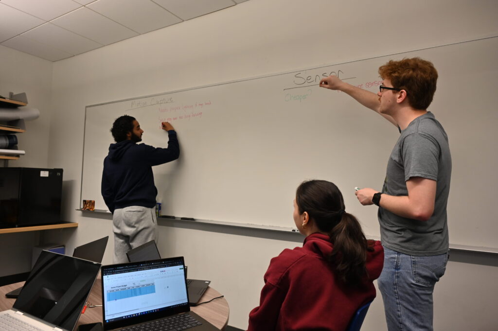 Jeremy pointing at the whiteboard while Hisham writes. Jasmine is at the table watching. The whiteboard shows the beginning stages for pros and cons of the Motion Capture and Sensor solutions for measuring hand angles.