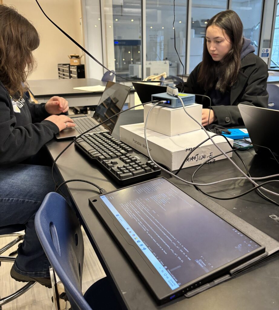 Stephanie and Jasmine working on their laptops with the mini PC and monitor used for the H.O.M.E. device.