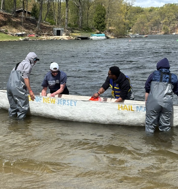The Hail Mary on the water during the swamp test. Team members (from left to right) Ethan Earley, Anthony Giorgi, Olivier Duverneau, and Rachel Alecci surround the canoe. Anthony and Olivier both hold orange buckets.
