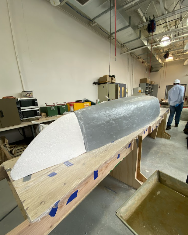 The fully prepared canoe mold sitting on a wooden table. Foam was shaped in order to make the ends of the canoe taper in the desired way. The center of the mold is wood that was covered in spackle and painted a dark gray.