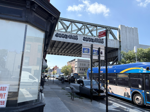 Image portrays congestion along 2nd Avenue during the midday peak hour. Multiple vehicles including a city bus, cars, and a van line the roadway beneath an elevated train track also depicted within the image. The busy area shows current traffic conditions at the site. Clear blue skies define the weather conditions.