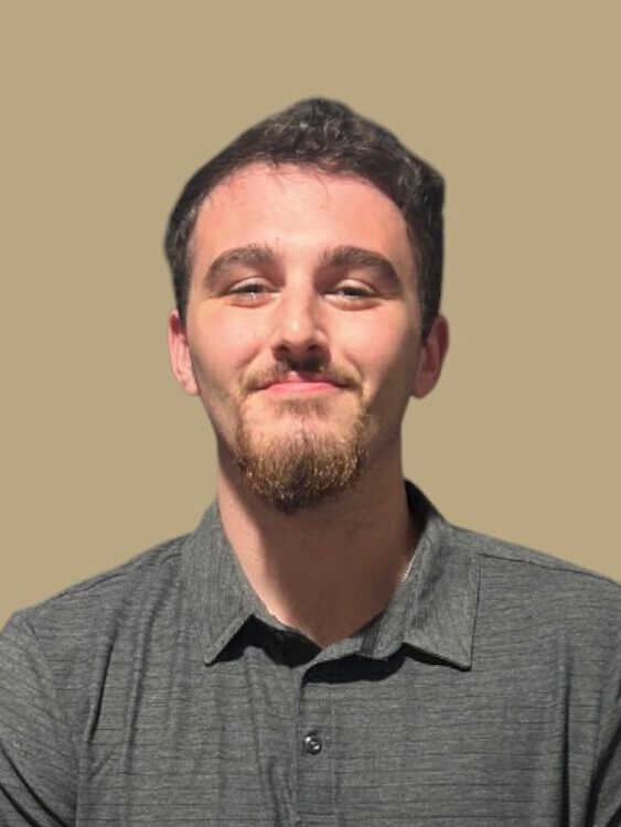 Young man with dark hair and facial hair, wearing a dark gray shirt against a beige background.