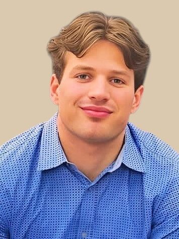 Young man with light brown hair, wearing a blue button-up shirt against a beige background.