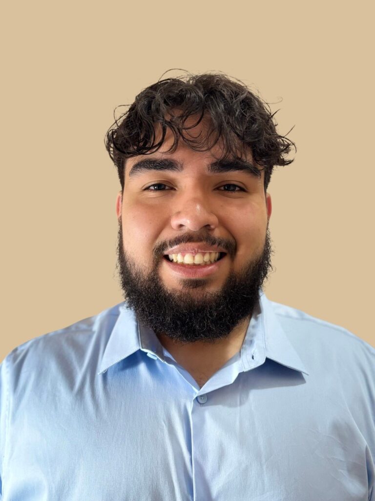 Young man with curly hair and facial hair, wearing a blue button-up shirt, against a beige background.