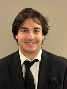 Young man with dark hair, wearing a black suit jacket, white shirt, and black tie against a beige background.