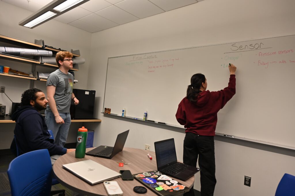 Jeremy and Hisham looking at the whiteboard while Jasmine is writing. The whiteboard shows the beginning stages for pros and cons of the Motion Capture and Sensor solutions for measuring hand angles.