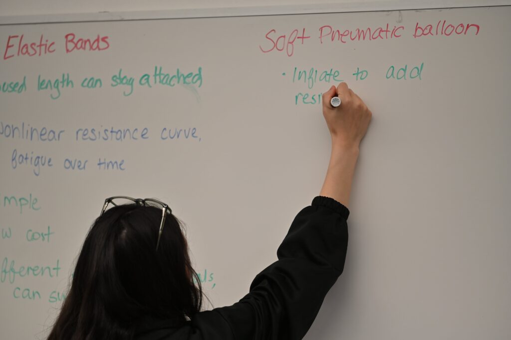 Close up of Jasmine writing "Inflate to add resistance" under the title "Soft Pneumatic balloon" on the whiteboard.