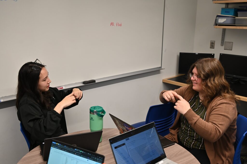 Jasmine and Stephanie sitting at the round table with index fingers overlapping. They are trying to understand how friction sliders would be implemented as resistive elements in the device.